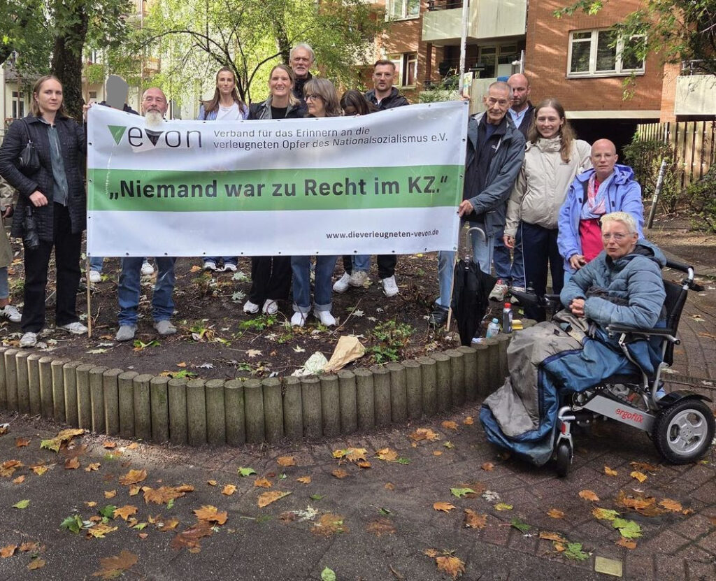 Eine Gruppe von Menschen steht gemeinsam hinter einem Banner des Vereins„Verband für das Erinnern an die verleugneten Opfer des Nationalsozialismus e. V.“. Auf dem Banner steht: „Niemand war zu Recht im KZ.“ 